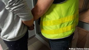 Zatrzymany skuty kajdankami trzymany od tyłu przez policjanta w żółtej kamizelce.
