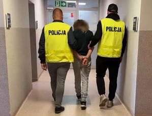 Policjanci w żółtych kamizelkach z napisem policja prowadzą zatrzymaną przez korytarz.