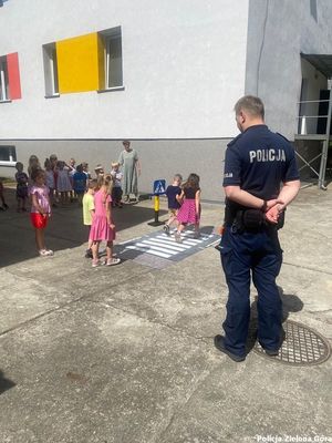 Policjant tłumaczy dzieciom jak w bezpieczny sposób przechodzić przez przejście dla pieszych.