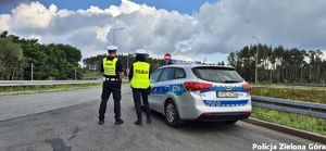 Policjantka i policjant ruchu drogowego, w żółtych kamizelkach z napisem Policja stoją koło radiowozu na drodze wylotowej z miasta.