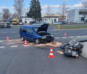 Zdjęcie przedstawia niebieski samochód osobowy uszkodzony z przodu po zderzeniu z motocyklem. Jego maska jest pogięta, uniesiona. Przed samochodem leży uszkodzony motocykl. Wokół pojazdów rozstawione są 4 pachołki.