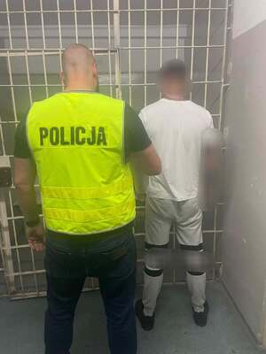 zdjęcie przedstawia stojącego w komisariacie przed kratą zatrzymanego mężczyznę w kajdankach. Za nim stoi policjant w kamizelce odblaskowej z napisem "Policja"